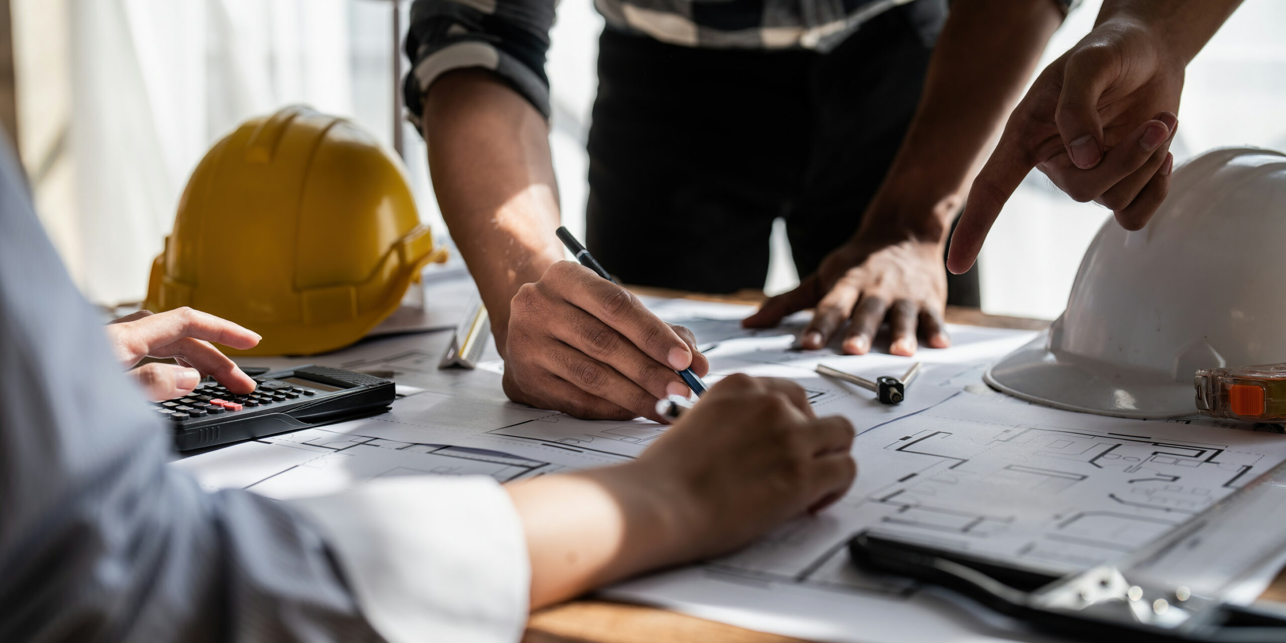 Three people collaborate over architectural blueprints on a desk, using pens and a calculator. Two safety helmets, one yellow and one white, sit nearby, indicating a construction or engineering project meeting.