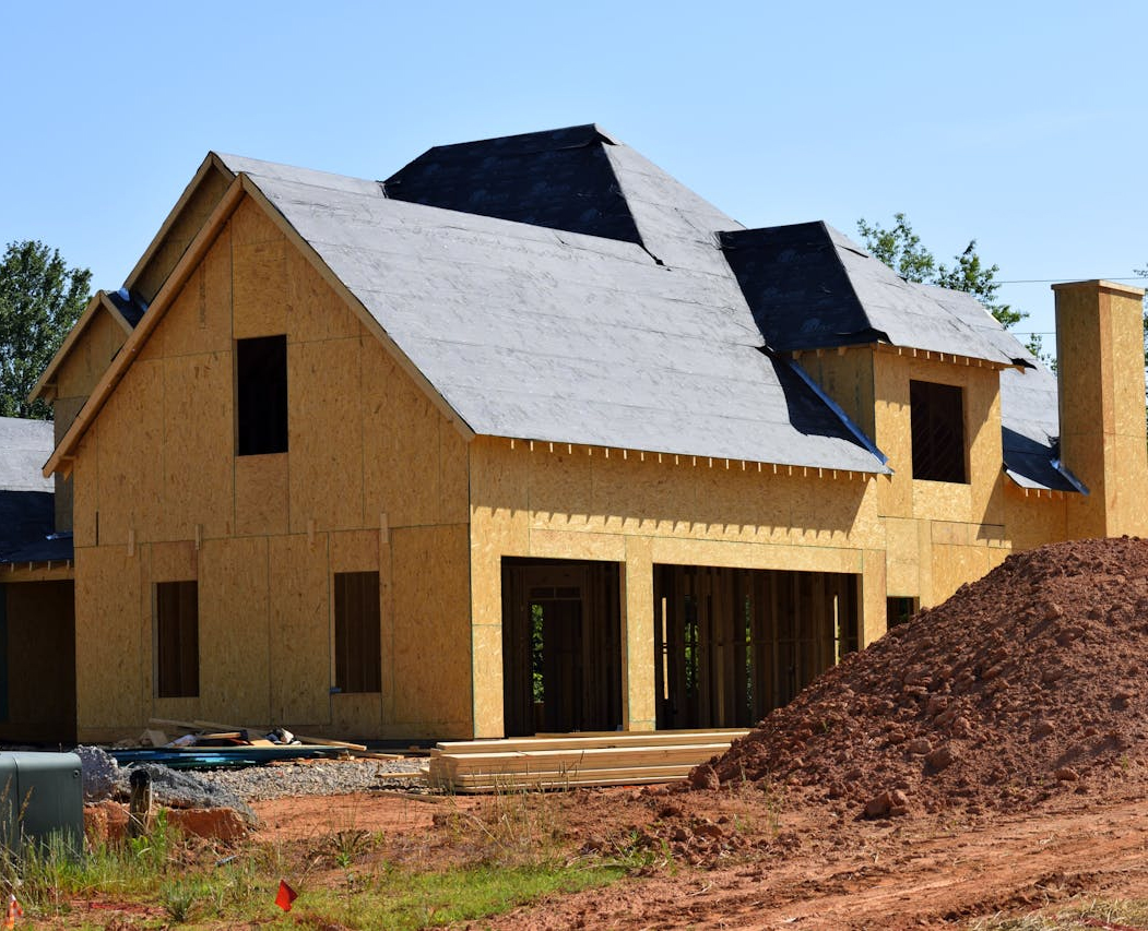 A large house under construction with exposed wooden framing and a black roofing underlayment, surrounded by dirt and building materials on a sunny day.