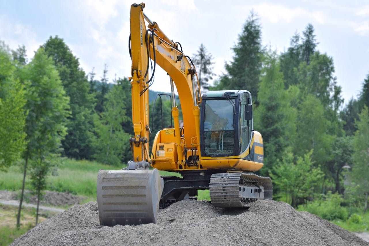 A yellow excavator with a large bucket sits on top of a gravel mound, surrounded by green trees and foliage under a partly cloudy sky.