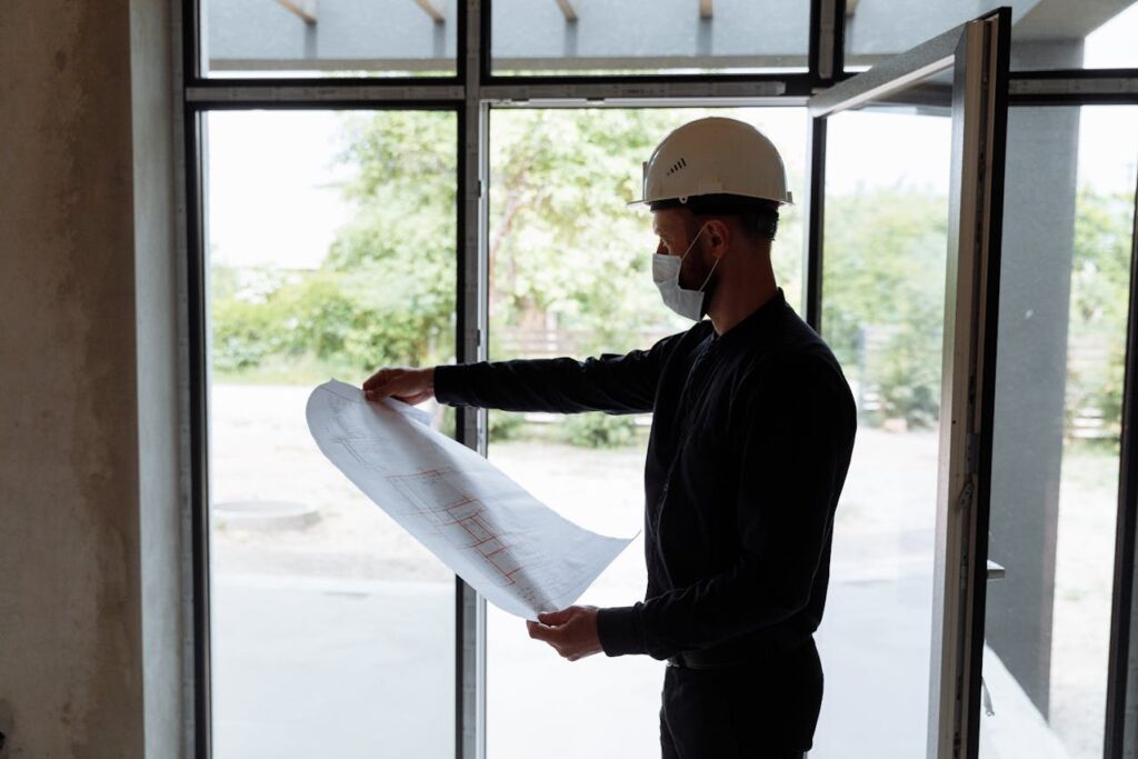A person wearing a white hard hat and a face mask stands indoors by an open door, holding and examining a large sheet of architectural blueprints. Natural light comes in through the glass windows behind them.