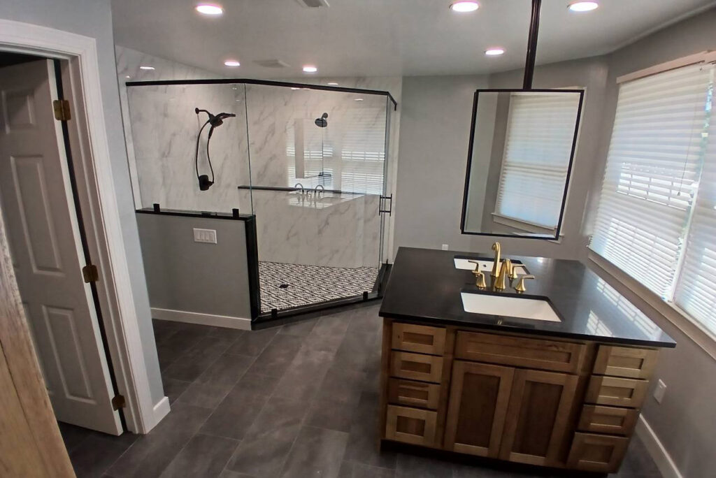 Modern bathroom with a glass-enclosed shower, black and white tile floor, single vanity with a dark countertop, gold faucet, and large mirror; windows with blinds let in natural light.