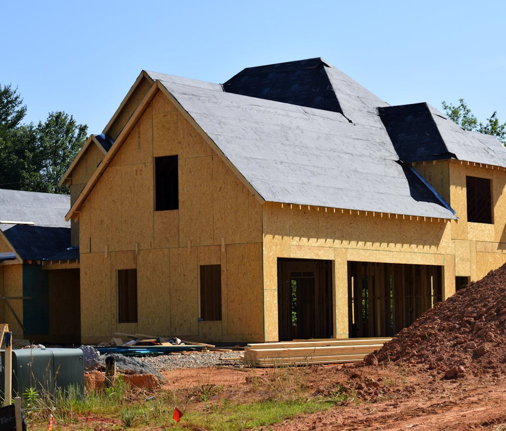 A house under construction with wooden framing and unfinished exterior walls, surrounded by dirt and construction materials, under a clear blue sky.