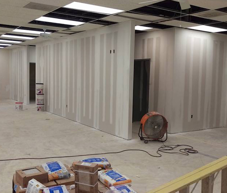 An unfinished interior room with bare drywall walls, some exposed ceiling panels, a large floor fan, paint cans, and construction materials scattered on the floor.