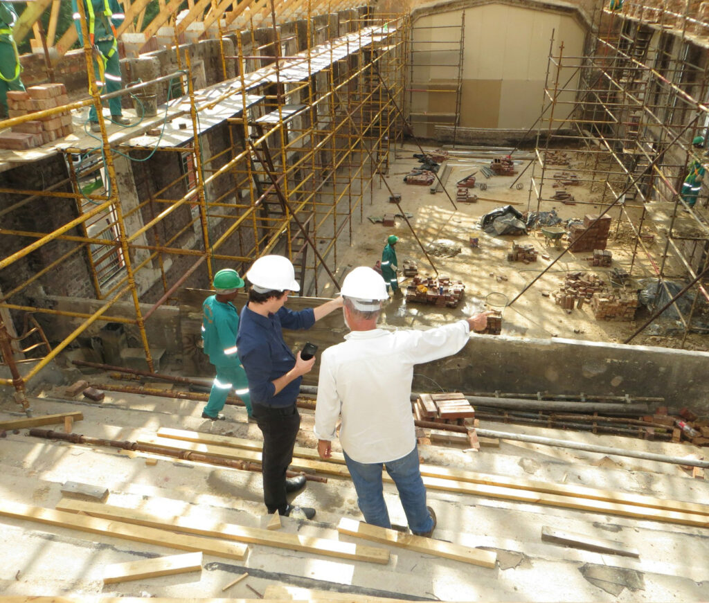 Two people wearing white hard hats stand on a construction site, discussing plans. Workers in green uniforms and helmets are nearby, with scaffolding, wooden beams, and building materials surrounding them.
