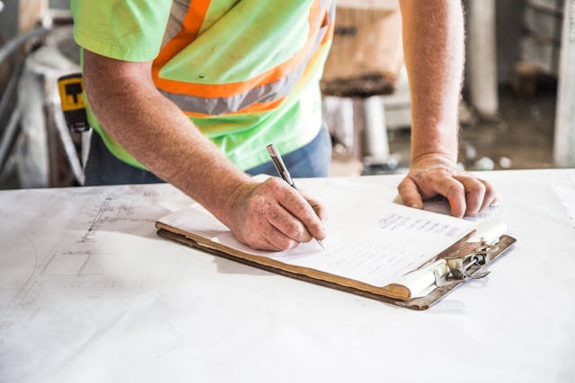 A person wearing a neon green safety shirt with orange reflective stripes writes on a clipboard with papers, leaning on a table covered with documents at a construction site.