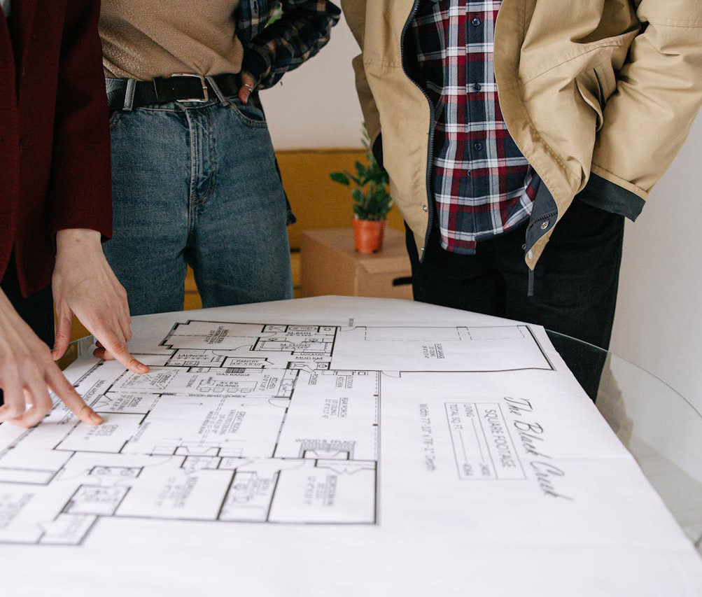 Three people stand around a table looking at and pointing to a large architectural floor plan blueprint, discussing details. A small plant and a brown box are visible in the background.