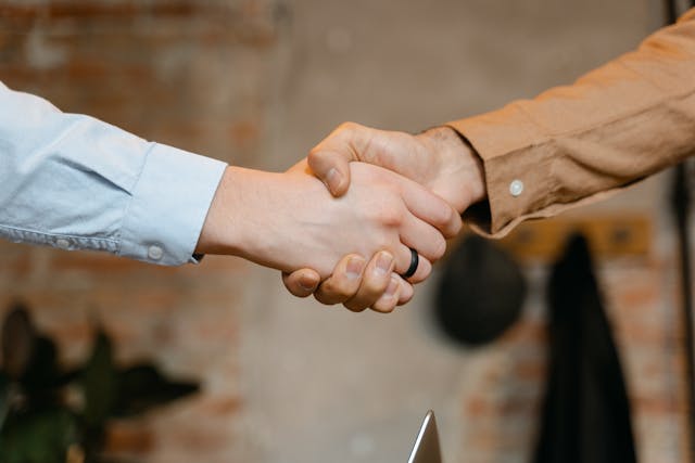 Two people wearing long-sleeved shirts are shaking hands, symbolizing agreement or partnership, with a blurred indoor background.