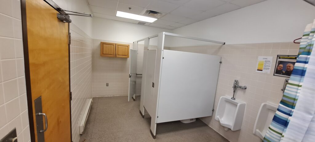 A clean, tiled men’s restroom with a wooden door, two stalls with white partitions, a urinal, a sink with a blue and white shower curtain, wall cabinets, and notices posted on the wall.