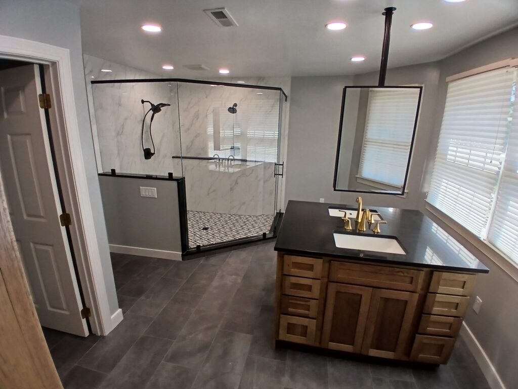 Modern bathroom with a glass-enclosed shower, gray tile floor, wooden vanity with black countertop, gold faucet, large mirror, and windows with blinds letting in natural light.